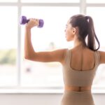 Modern açık bej dar spor giysili genç kadının mavi dambıl tutarken ve fit vücudunu gösterirken arkadan görünümü